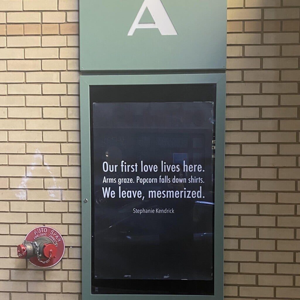 A framed poster of a haiku by Stephanie Kendrick, for the Athena Cinema's annual haiku contest. White text on black background, it reads: 
"Our first love lives here.
Arms graze. Popcorn falls down shirts.
We leave, mesmerized."
