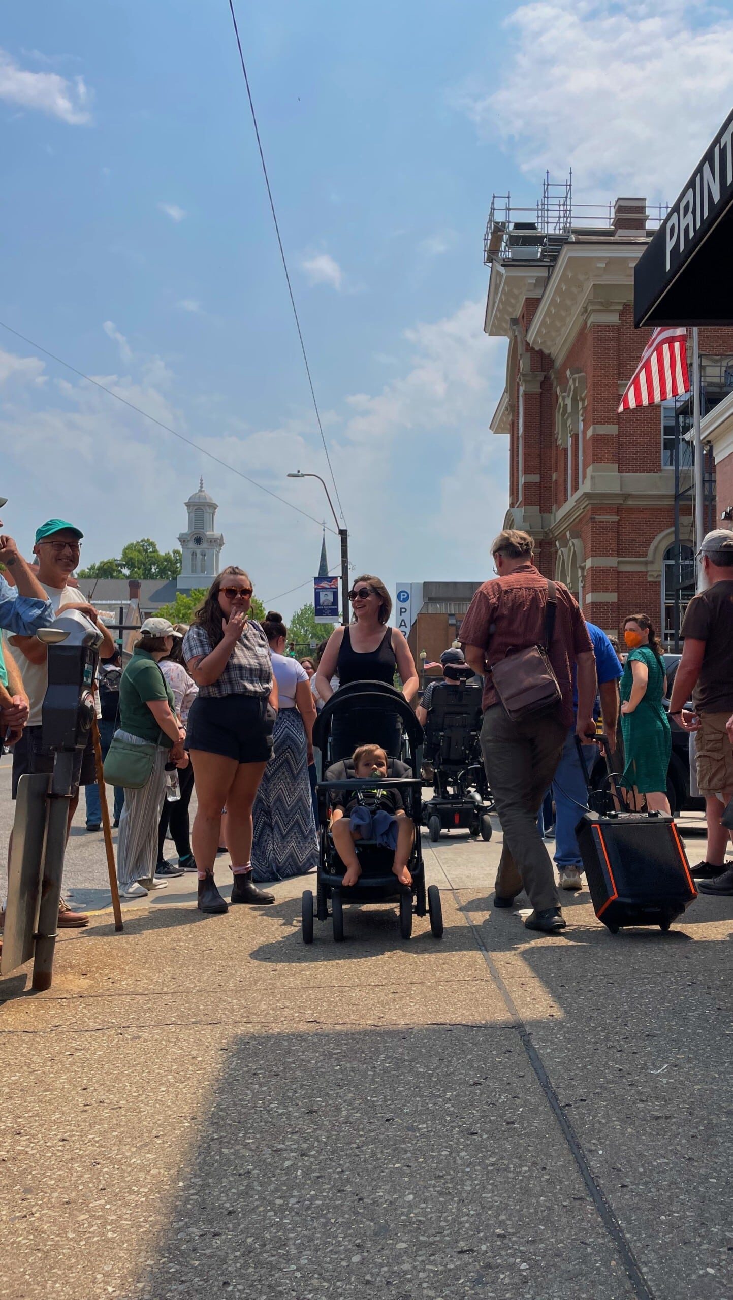 A crowd moves along the sidewalk on a summer day. In the center of the photo, a person pushes a stroller.