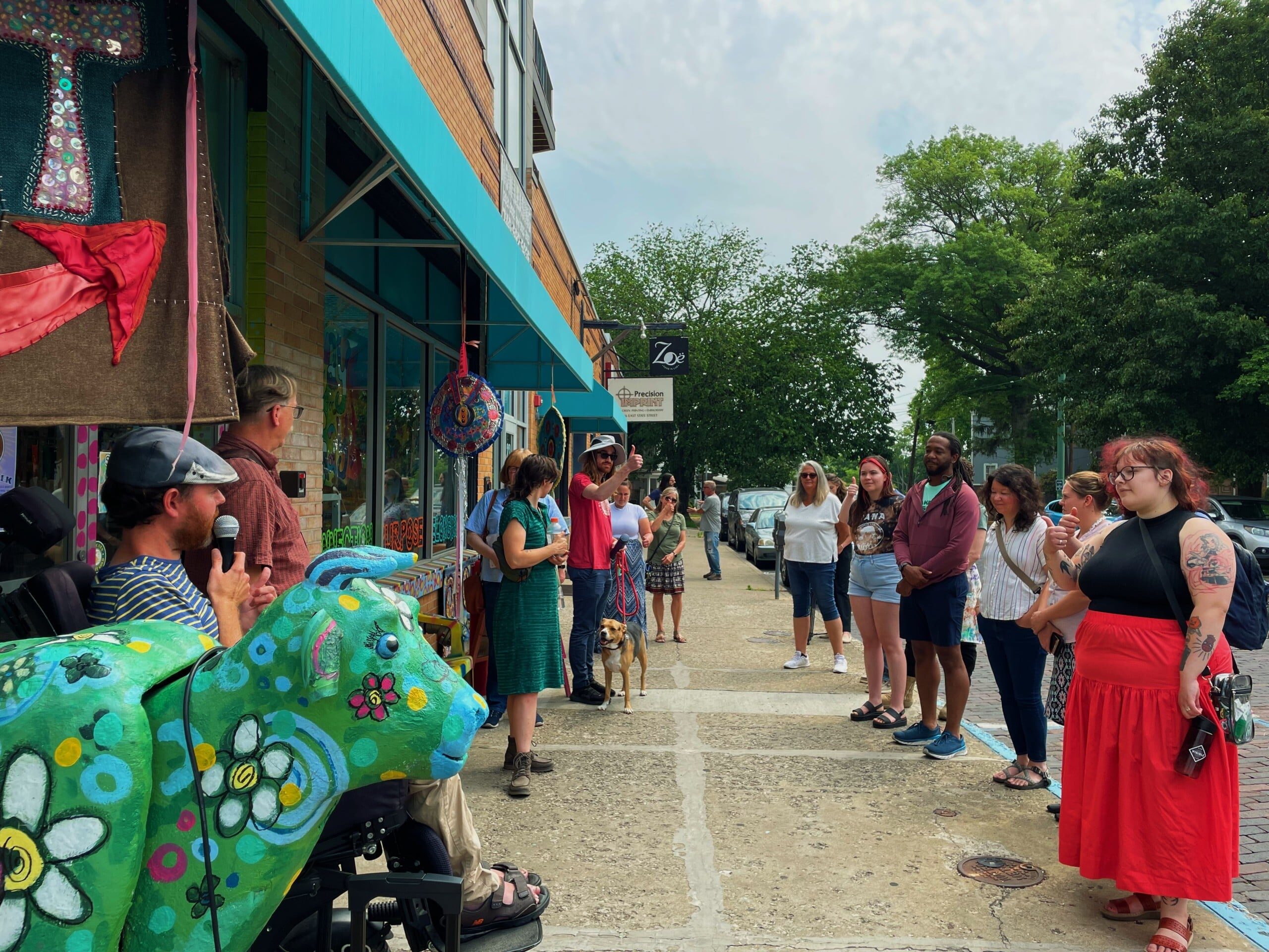 A person using a wheelchair holds a microphone and addresses a crowd gathered along the sidewalk outside a storefront. Colorful artwork features in the foreground.