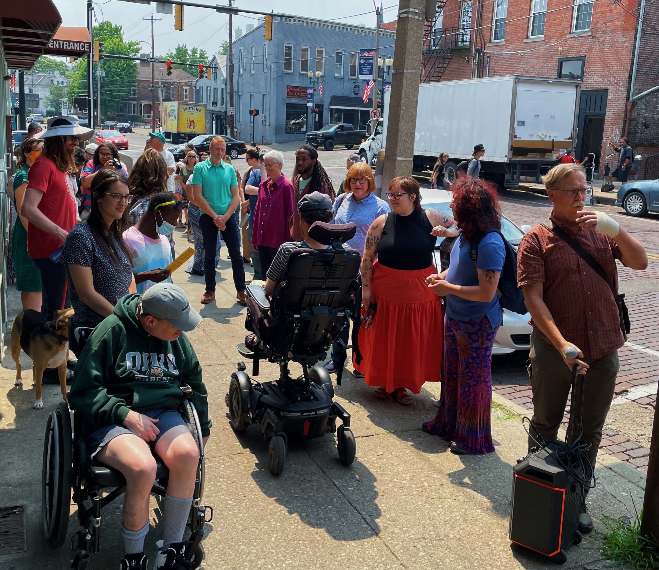 A crowd gathers on the sidewalk. At the center of the photo is a person using a wheelchair, addressing the mostly standing crowd.