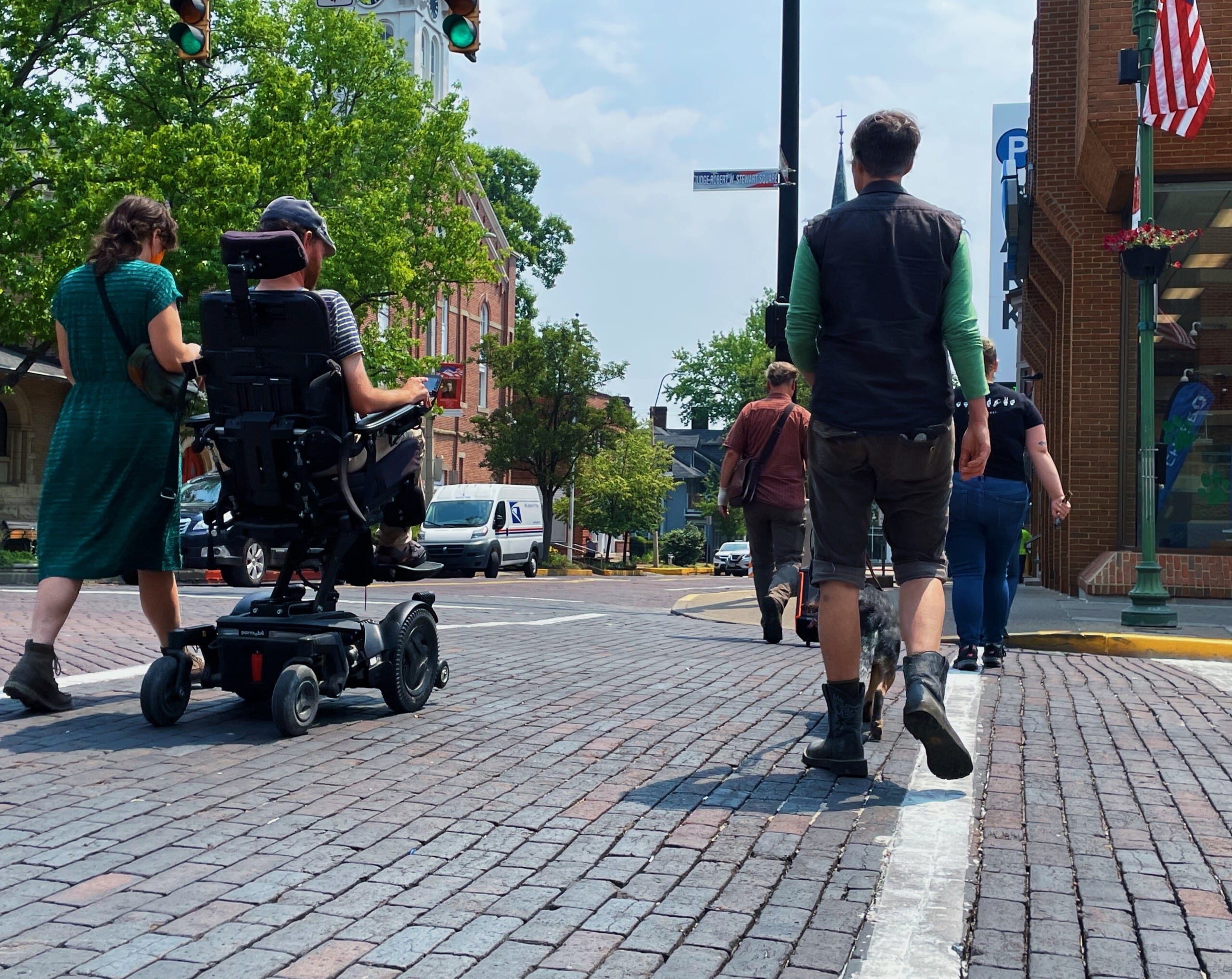 A group crosses a brick crosswalk on a warm sunny day. One person in the crowd uses a wheelchair.