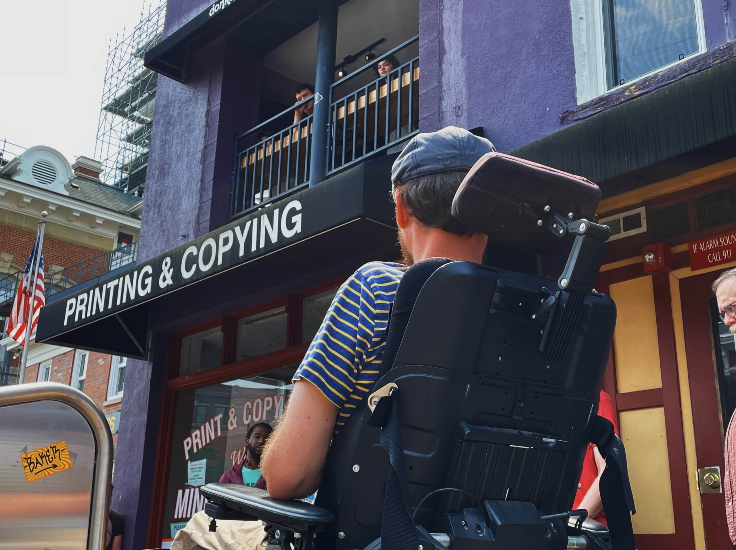 A man in a wheelchair, pictured from behind, looks at a printing and copying storefront.