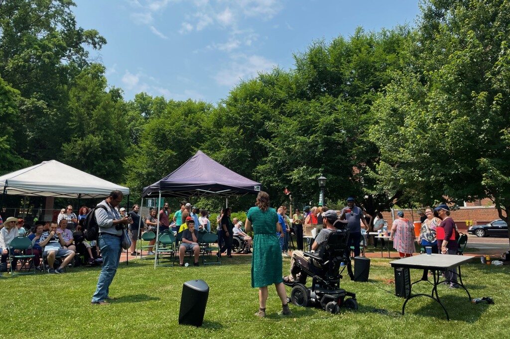 On a sunny day, a crowd gathers on mowed grass with trees surrounding the group on two sides. Multiple tents positioned throughout the crowd offer shade to attendees. A person using a wheelchair and another person standing address the crowd, both pictured from behind.