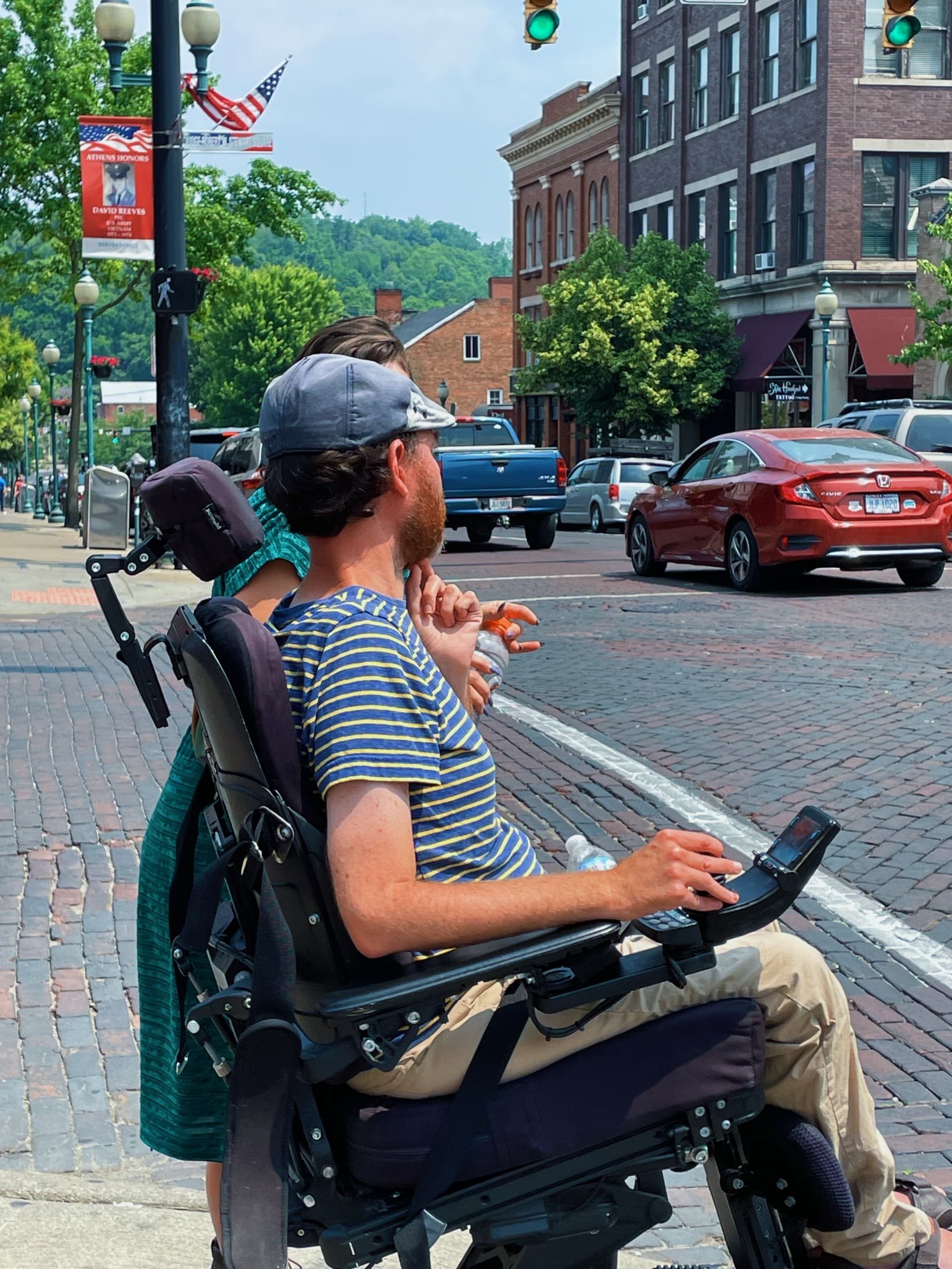 A person crosses a brick street using a wheelchair on a sunny day. Photo shows a side view of the individual. Cars drive along the street in the background.