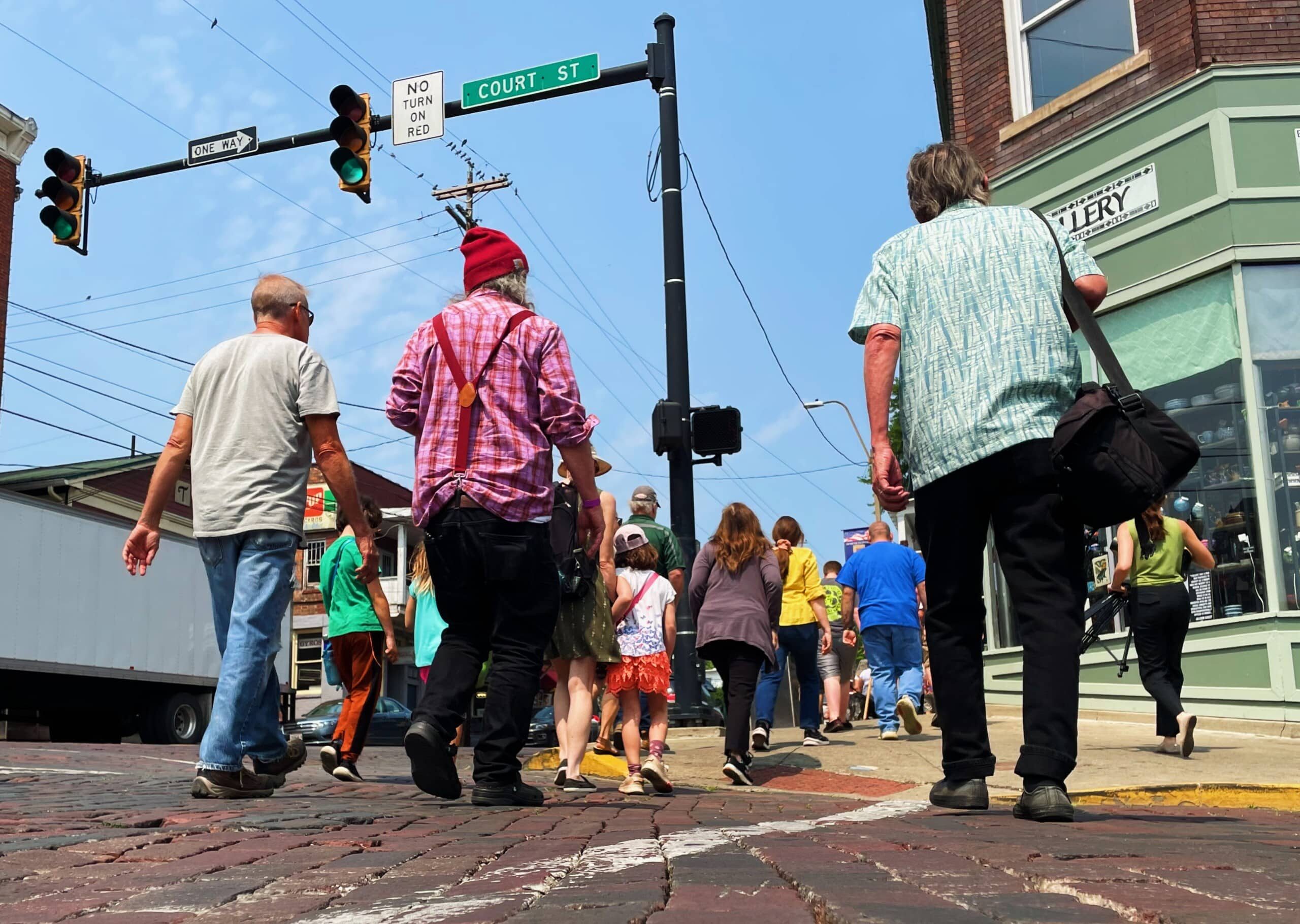 A crowd walks across a brick crosswalk toward a storefront on a summer day under a bright blue sky.