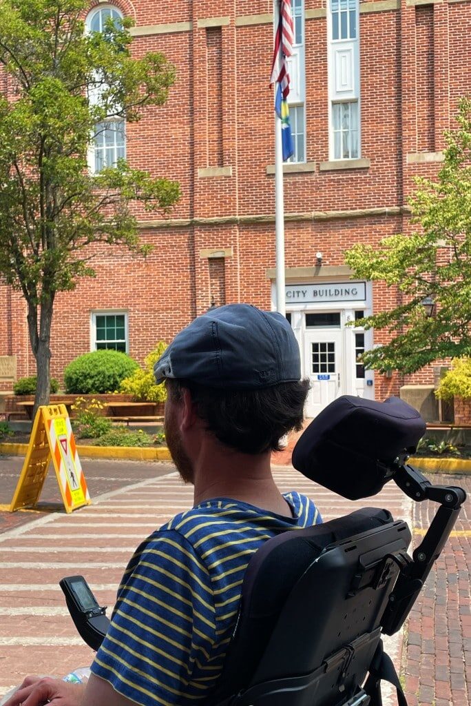 A person using a wheelchair approaches an intersection that features a crosswalk with a stamped brick appearance. The person in the foreground is shaded, while the background is brightly lit by the sun.