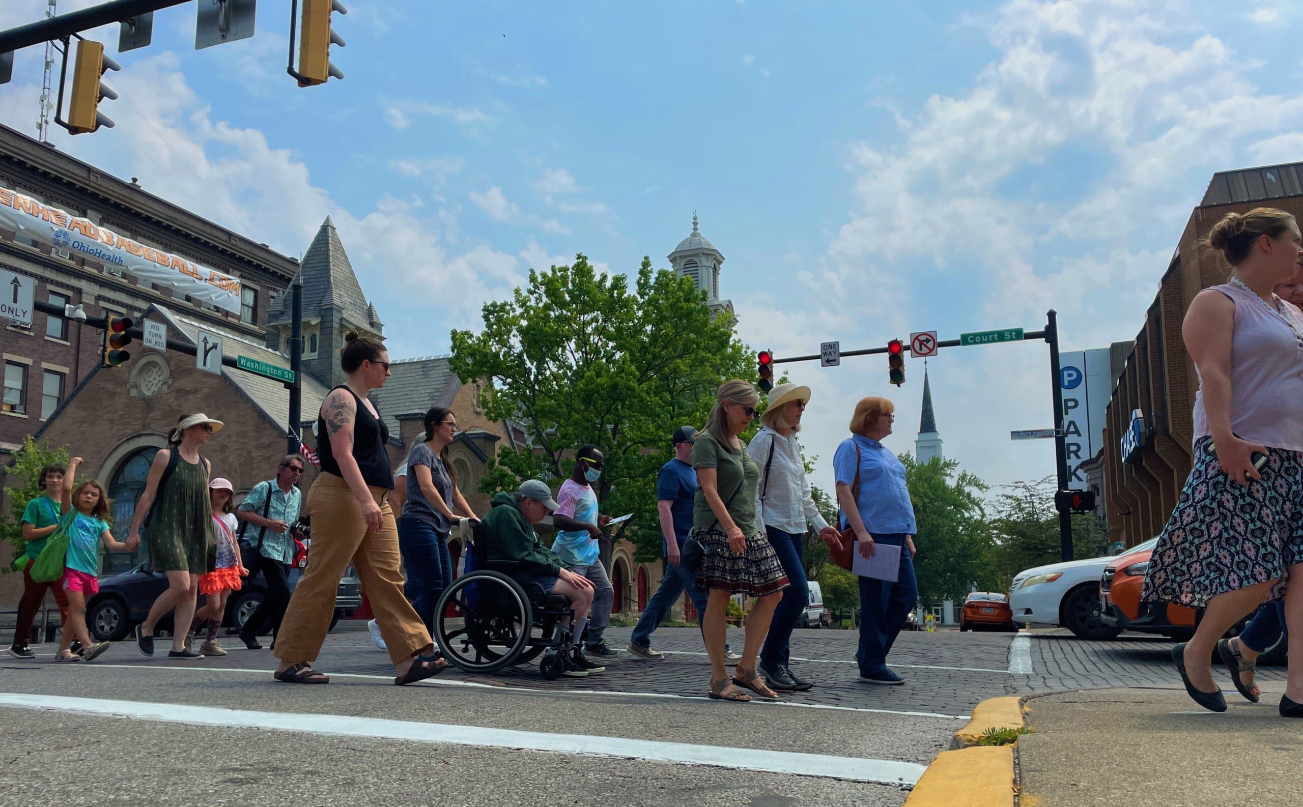A crowd crosses an intersection via a crosswalk on a sunny summer day.