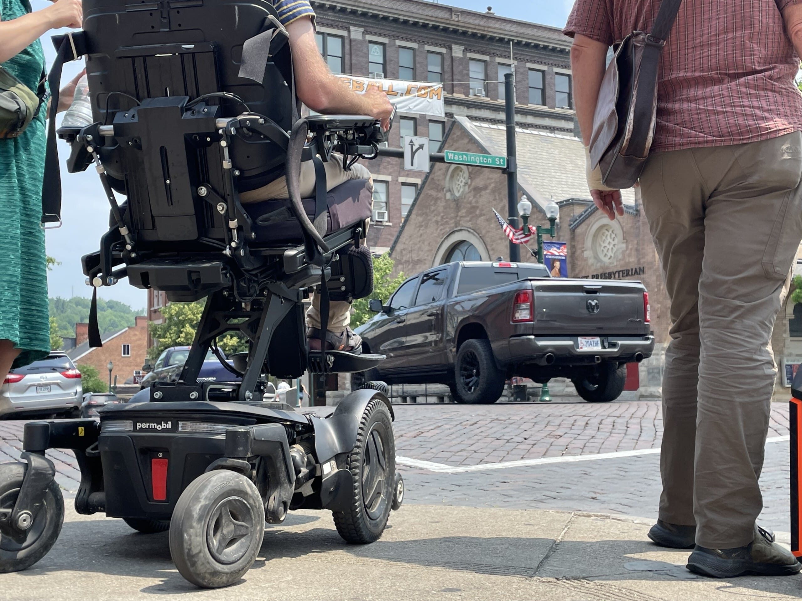 The people cross a brick street, one of whom uses a wheelchair. A truck passes through the middle of the intersection. Photo is taken from ground-level.