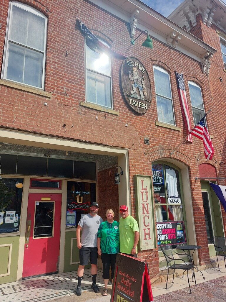 Three family members stand with their arms over one another in front of The Mine Tavern's storefront.