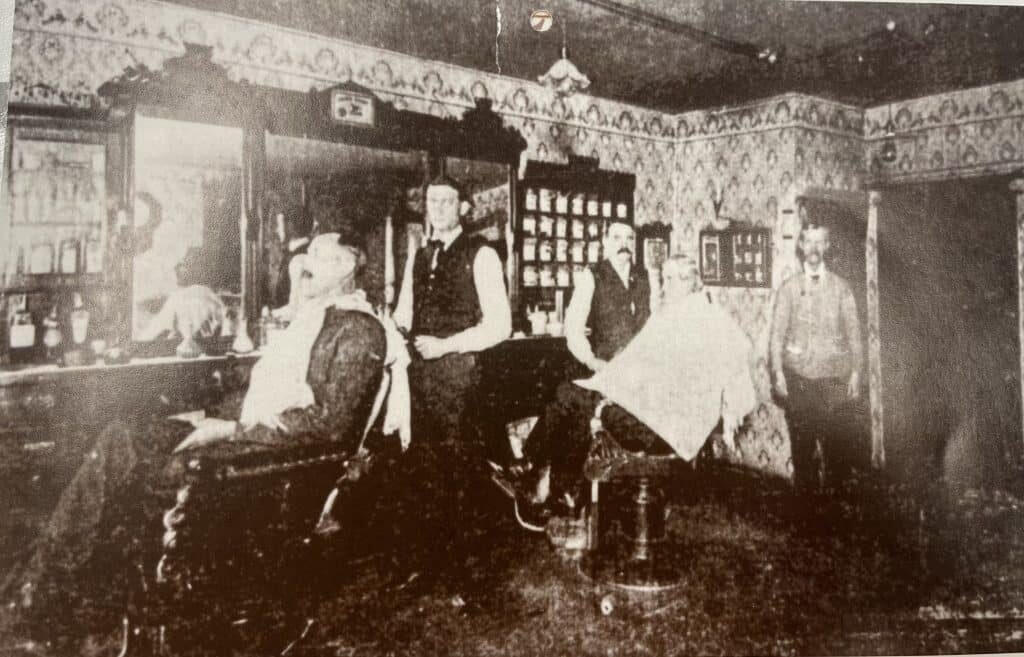 Grainy, black-and-white image of two patrons seated in a barber shop, with two barbers posted beside them, an another individual standing to the side.