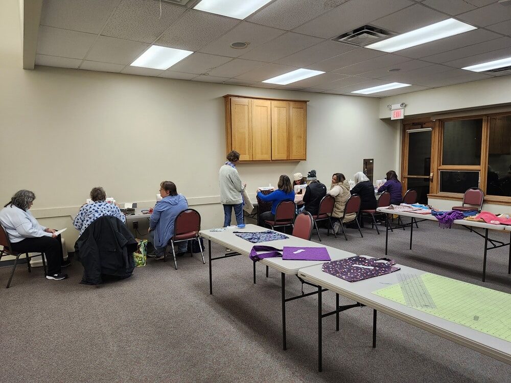 People sit and sew together at tables in a conference room.
