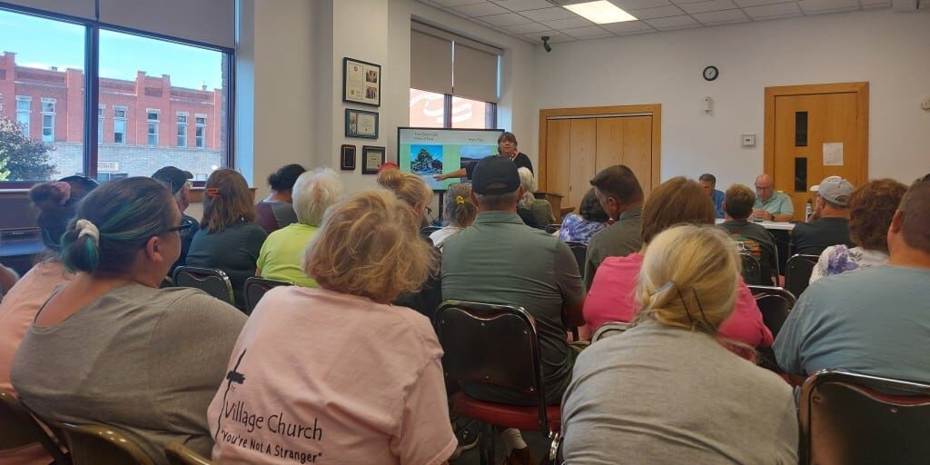 Numerous people sit before a woman gesturing to her presentation.