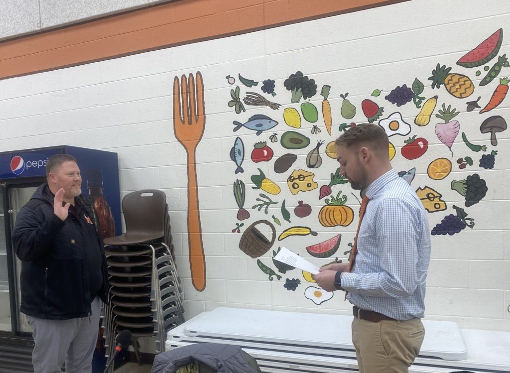 Two men stand, one with his hand raised as the other reads an oath from paper.