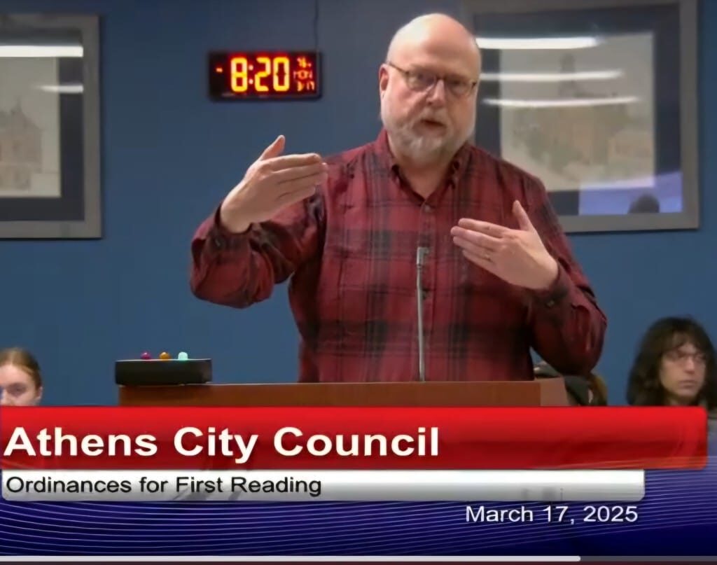 A man in a red flannel shirt speaks behind a podium. 