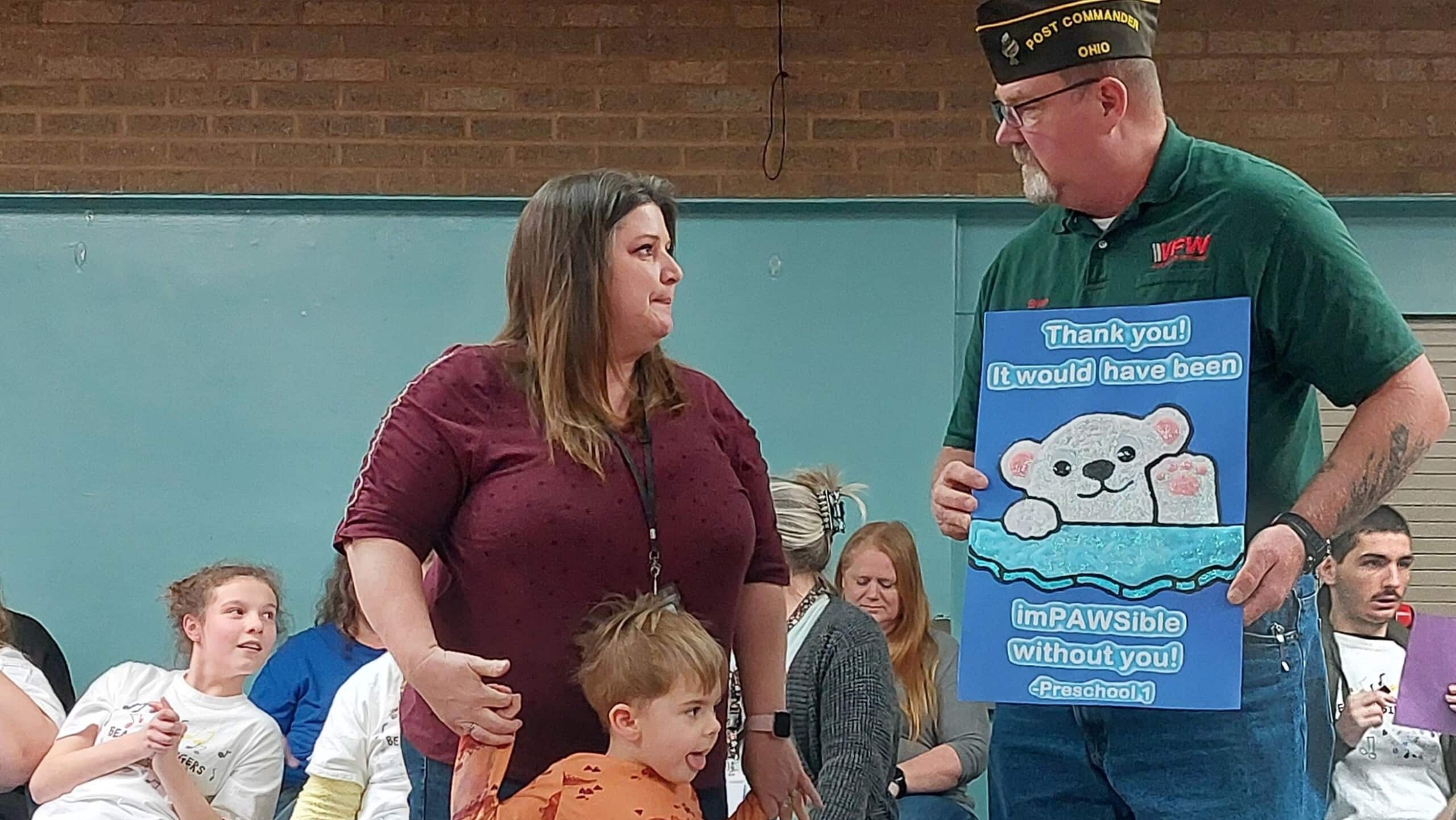 A Beacon School student and staff member present Brian Johnson, commander of the Albany VFW Post 9893, with a card. The student is a standing before his teacher. Johnson holds the card, that features a cartoon polar bear against blue background, that reads: "Thank you! it would have been impAWSible without you" - Preschool, 1 (referring to the student and staff). The polar bear gestures with its paw up, like a wave.