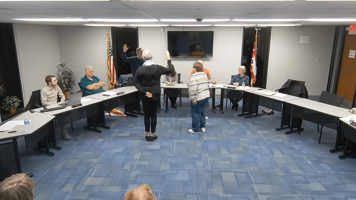 Two women stand in the foreground raising their right hands, taking the oath of office. Behind them are council members, seated, at a desk reaching across the room.