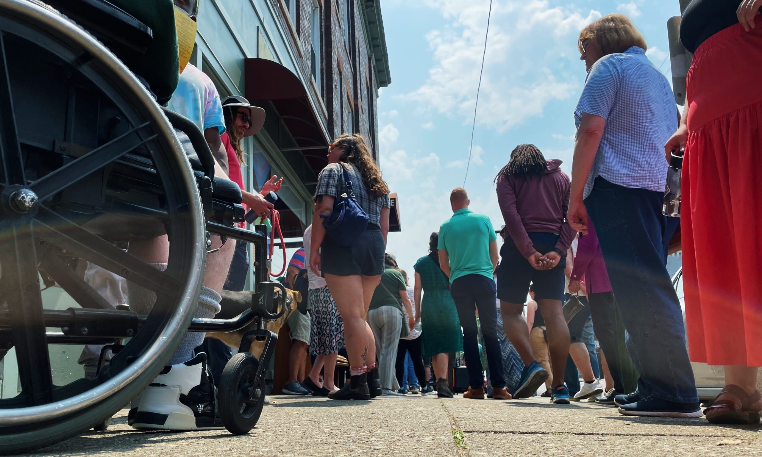 A crowd gathers on the sidewalk in Athens. Photo taken from the ground shows the wheel of a mobility device.
