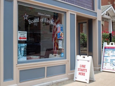 The exterior of the Athens County Board of Elections Office, featuring a large glass window with the office's name printed across it. An American flag is visible behind the glass. In front of the building is a sign that reads, "Line starts here."