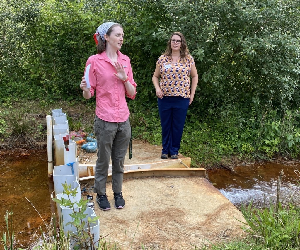 Two people stand on a small wooden bridge over a bright orange stream, one gesturing while speaking.