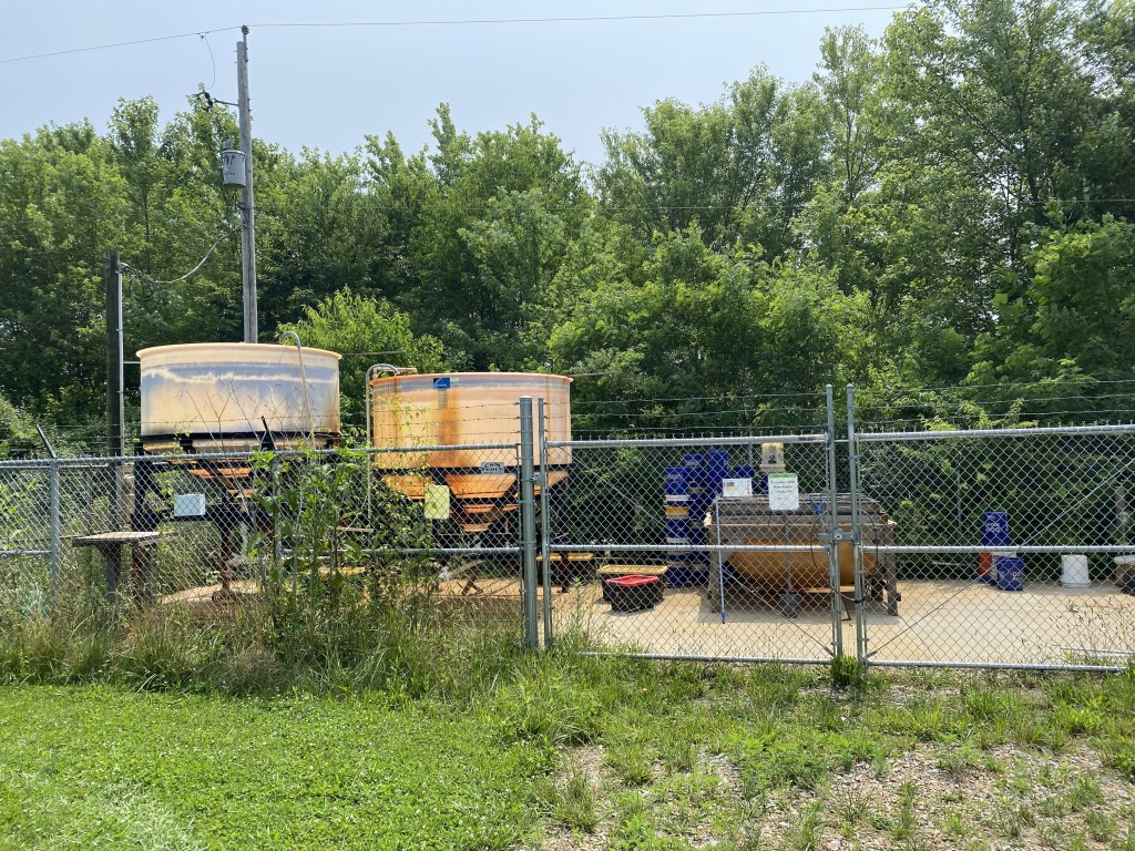 A fenced area featuring industrial equipment sits along a lush green tree-line.
