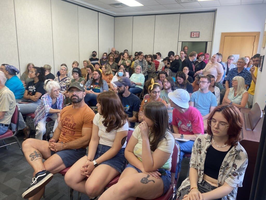An overflow crowd, dozens of people, at the July 19 board meeting. People of all ages sit, stand and talk to one n=another.