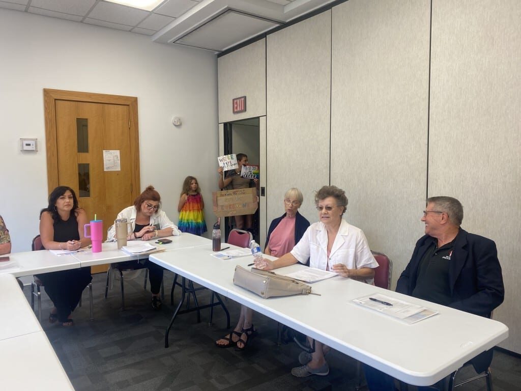 Five people sit at an L-shaped conference table with demonstrators, one in a rainbow dress and another holding signs, stand in a doorway.