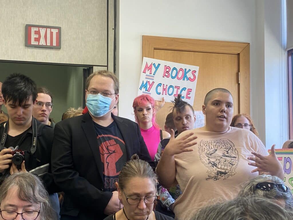 A crowded room full of demonstrators. A person in the front gestures, talking with their hands. A sign in the back reads, "MY BOOKS / MY CHOICE". Below each "MY" is the transgender flag. The bubble letters are in blue and pink, like the flag.