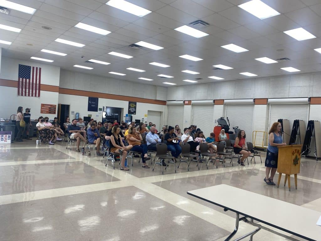 A woman at the forefront speaks at a podium while a large crowd sits in several rows of chairs behind her.