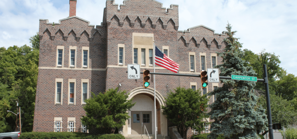 A photo of the Athens Armory in the summertime, an American flag waving in the forefront, lush trees surrounding it on either side.