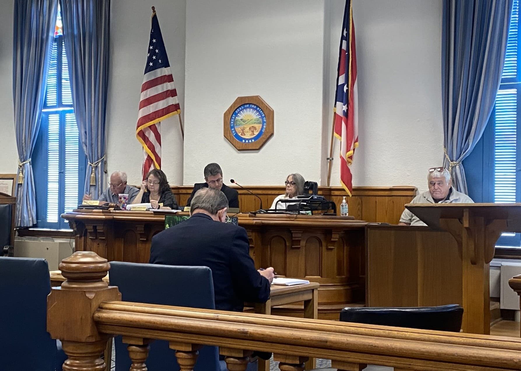 A panel of four sits at an elevated wooden table in a courtroom, with the American and Ohio flags and the Ohio seal behind them. A man sits at the witness stand, while an another sits in the foreground facing the panel and witness.