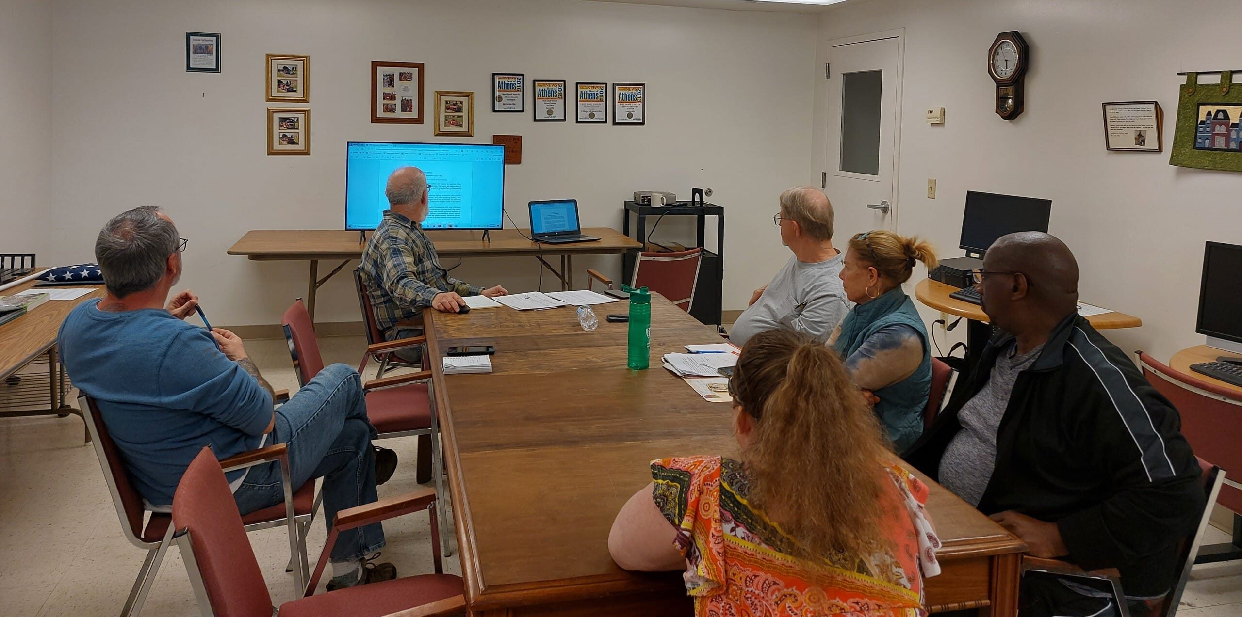 Six people sitting at a table in a conference room looking at a large screen.