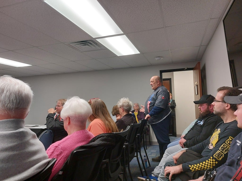 Behind a couple of rows of chairs, standing in a crowded room, Opha Lawson addresses the council.