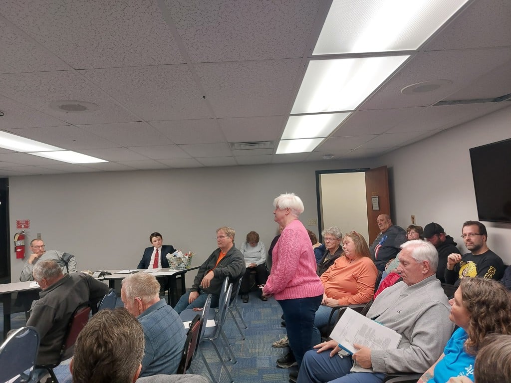 Sue Powell stands in the middle of a crowd, seated in rows of chairs.