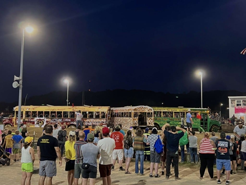 A large crowd stands in front of three school buses that are participating in the Athens County Fair's school bus demo derby.