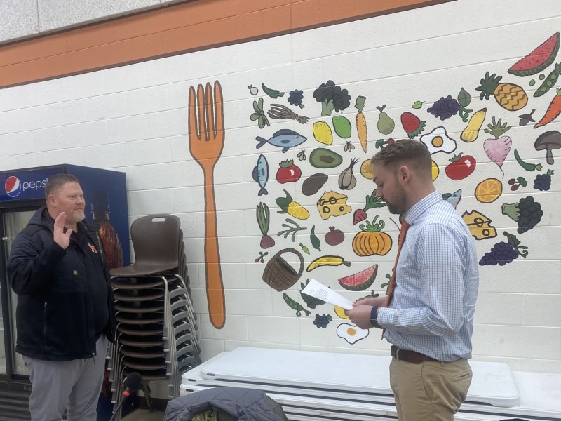 Two men stand, one with his hand raised as the other reads an oath from paper.
