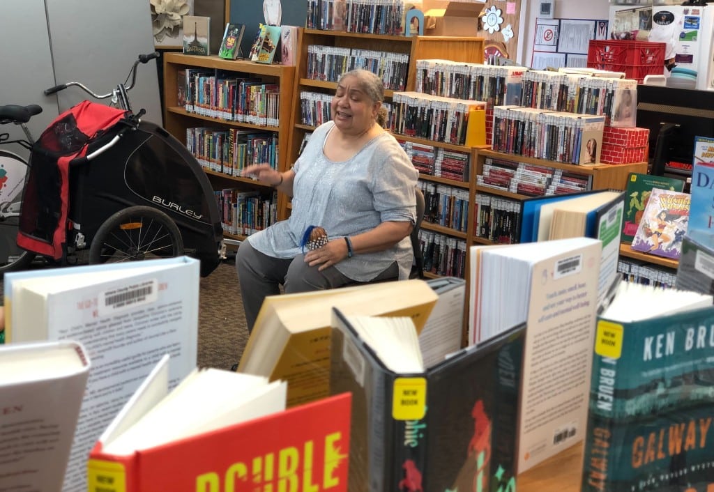 A woman, seated, speaks amongst shelves of library books.