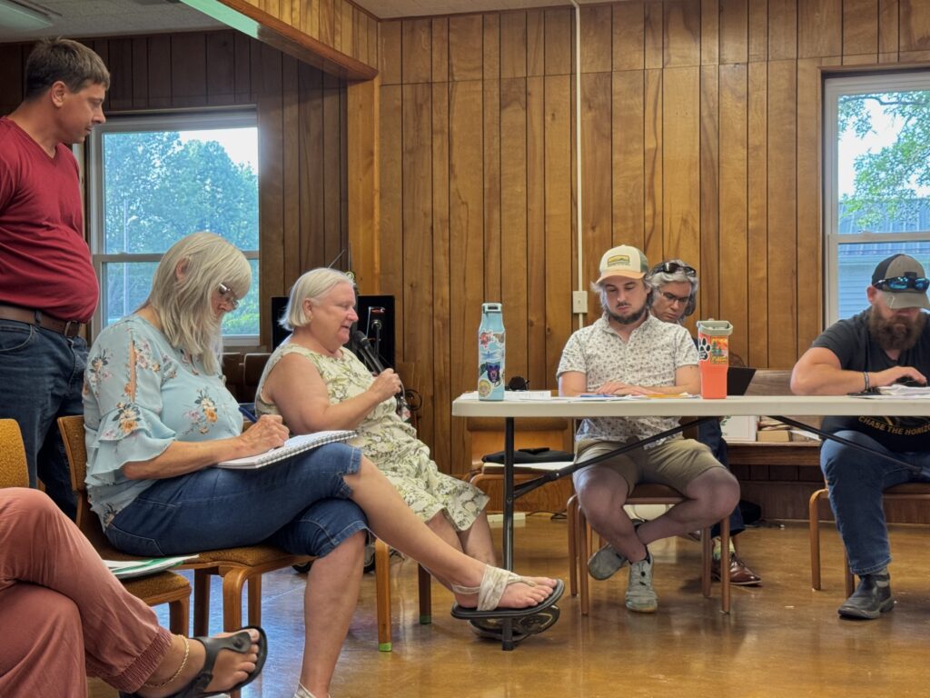 Group of people sitting, some behind a table. One person holding a microphone