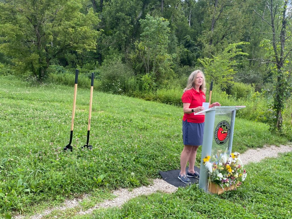 A woman in a red shirt and grey skit stands at a glass podium featuring the Ohio Department of Natural Resources logo. Behind her, in a field, shovels stand in the ground.