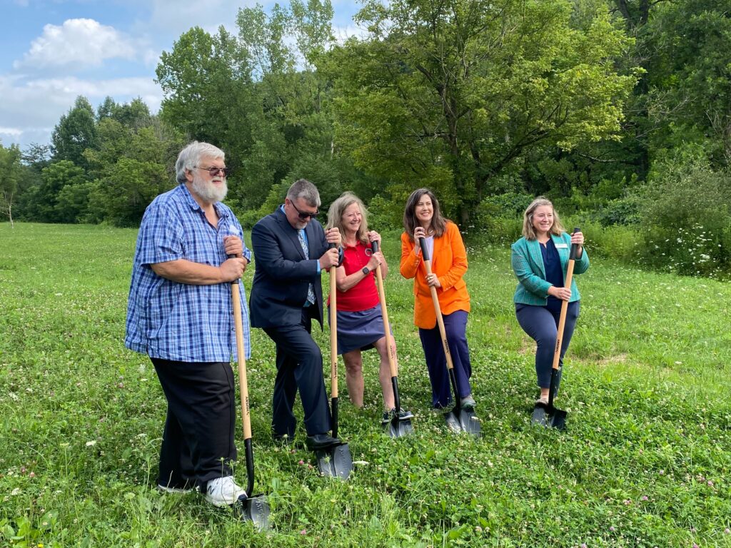 Five people push shovels into a field and pose for a photo, in front of a forest.