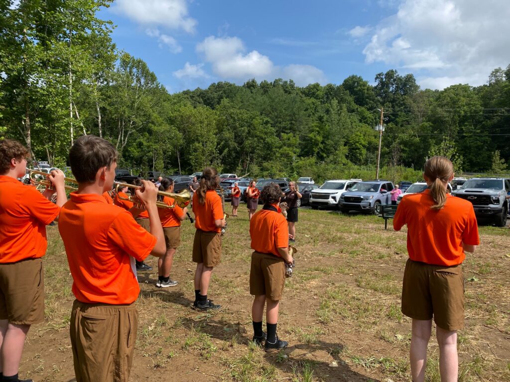 Two rows of uniformer performers, pictured from behind, play instruments. They stand on a field in front of their conductor and a parking lot.