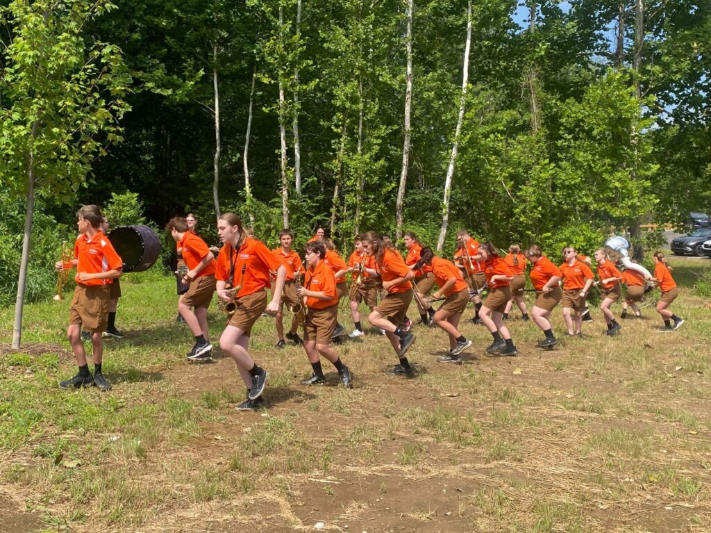 About 20 band performers in uniform turn in lock-step, holding their instruments and standing on dirt and grass in front of a forest.