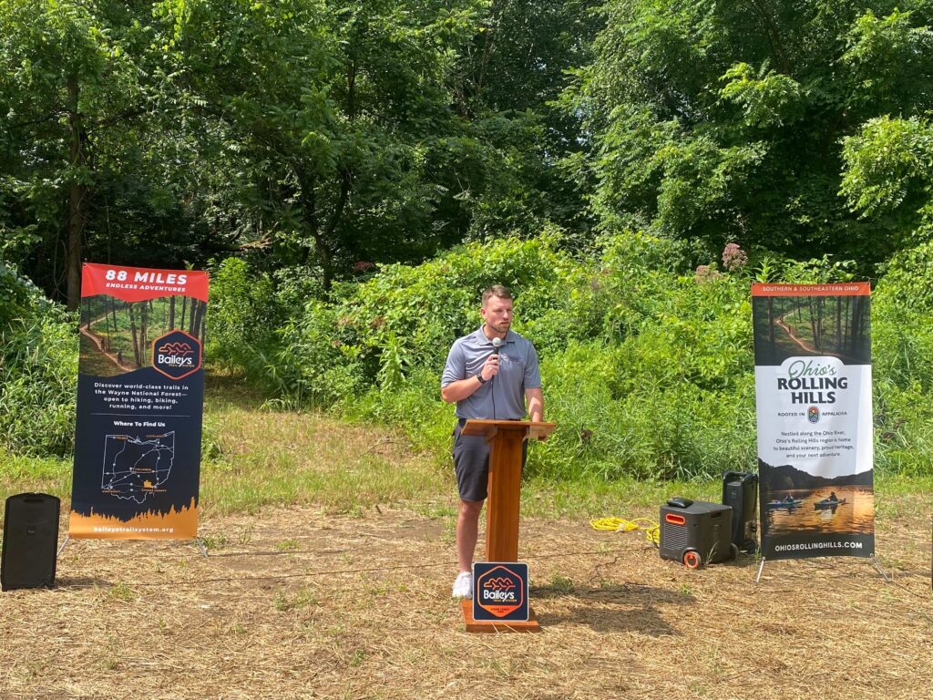 A man in a grey shirt and shorts holds a microphone at a podium, in front of two promotional banners that advertise the Baileys Trail System and a regional brand, Ohio's Rolling Hills. The man stands on dirt and grass in front of a forest.