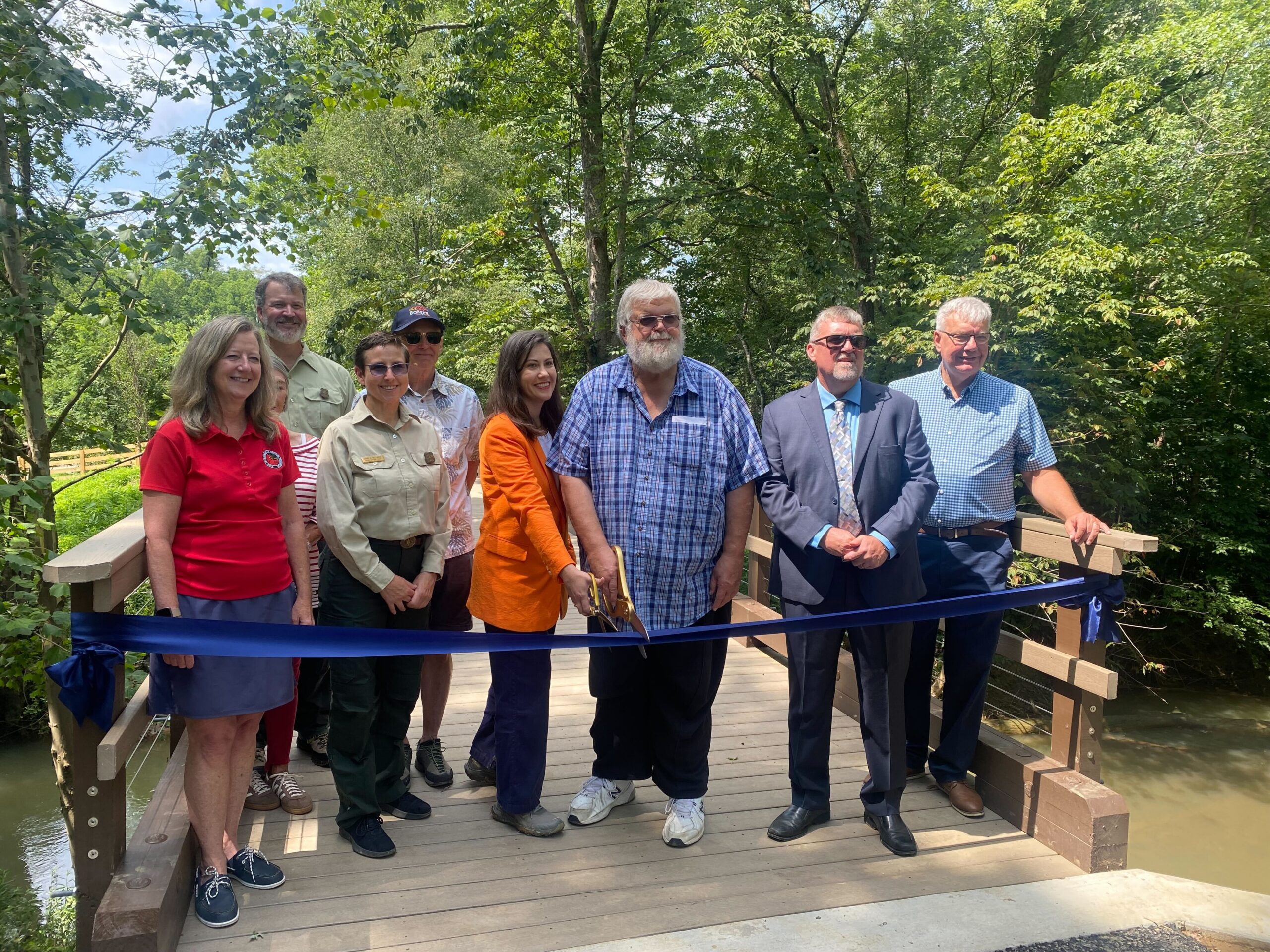 A group of nine people stands on a bridge that crosses a creek in the forest. A ribbon hangs in front of them, and the two people at the center hold large golden scissors to the ribbon.