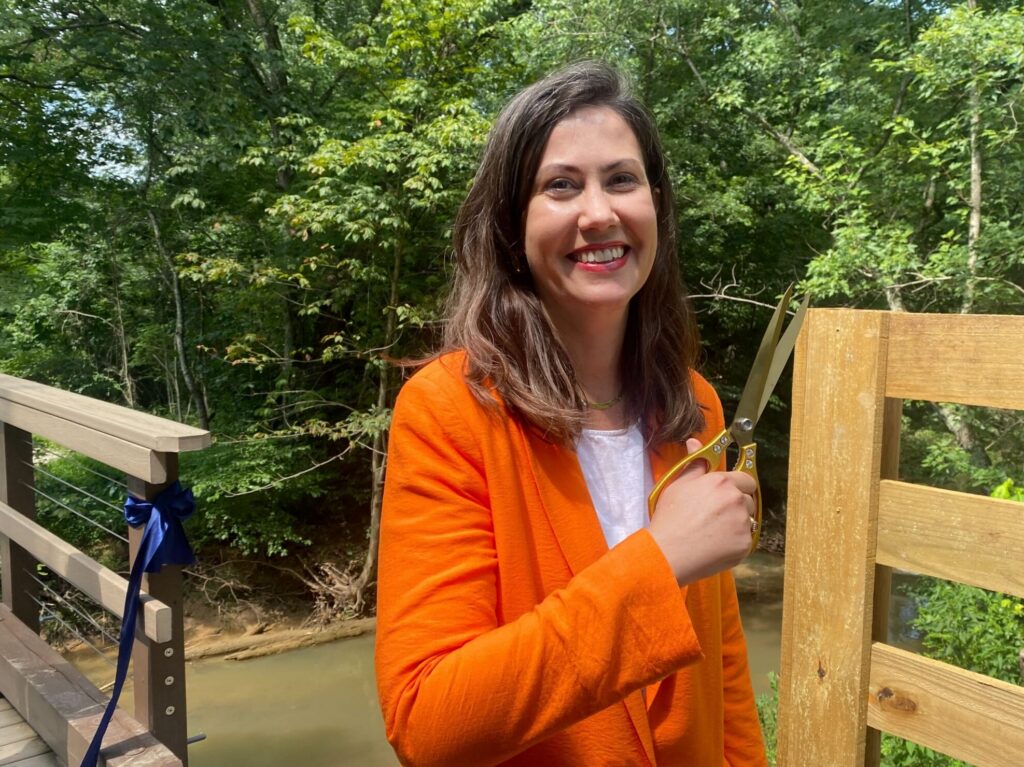 A woman in an orange blazer, pictured in front of a bridge that crosses a creek, holds a pair of golden scissors and smiles.