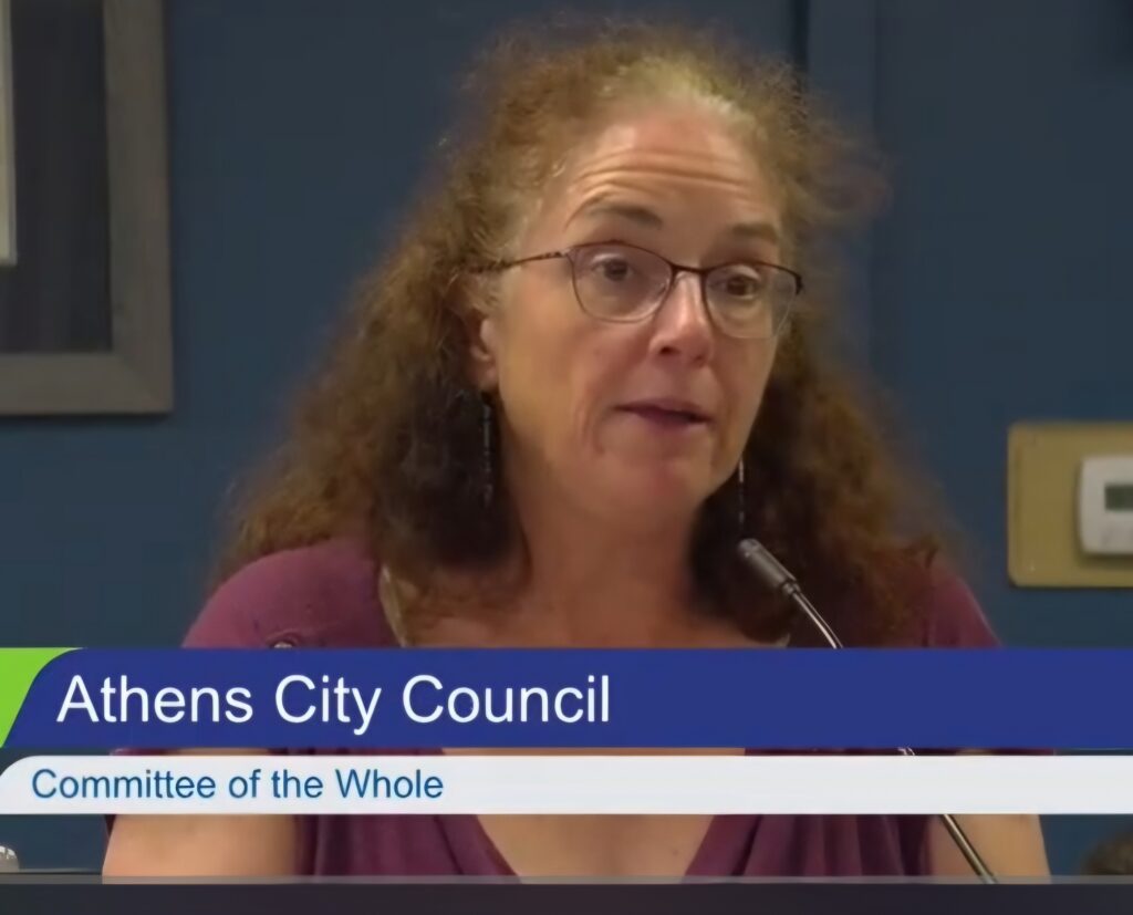 A woman speaks at a microphone. A banner reading "Athens City Council" overlays the image.