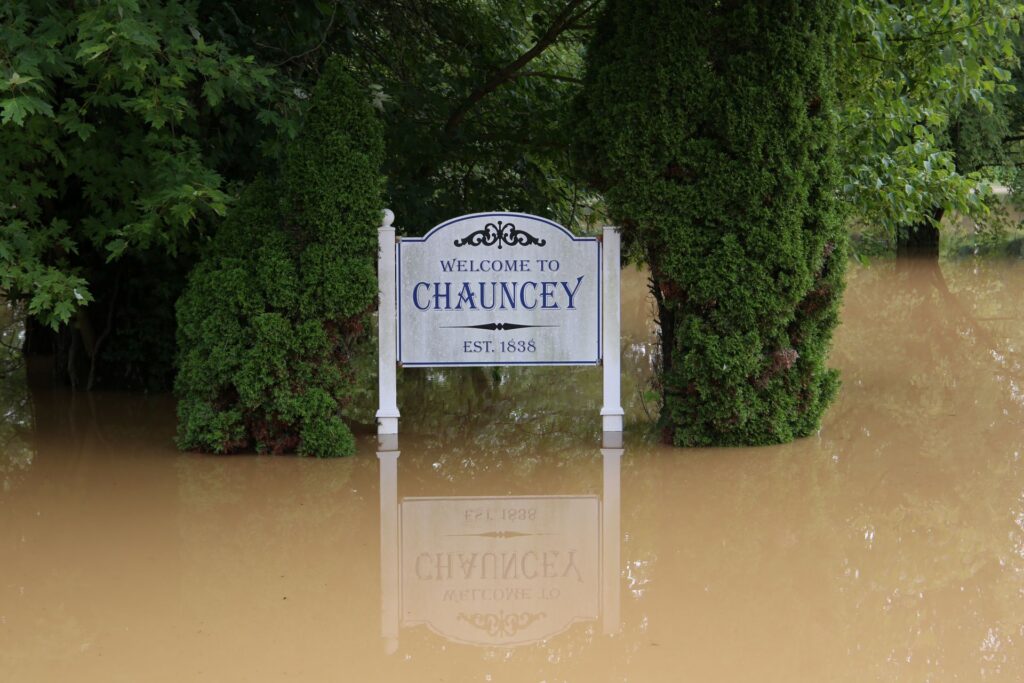 The "Welcome To Chauncey" sign is reflected by floodwater.