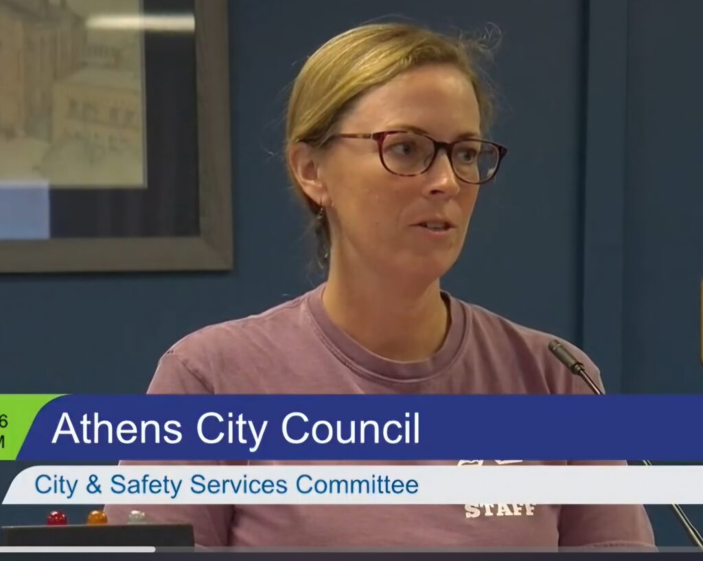 A woman speaks at a microphone. A banner reading "Athens City Council" overlays the image.