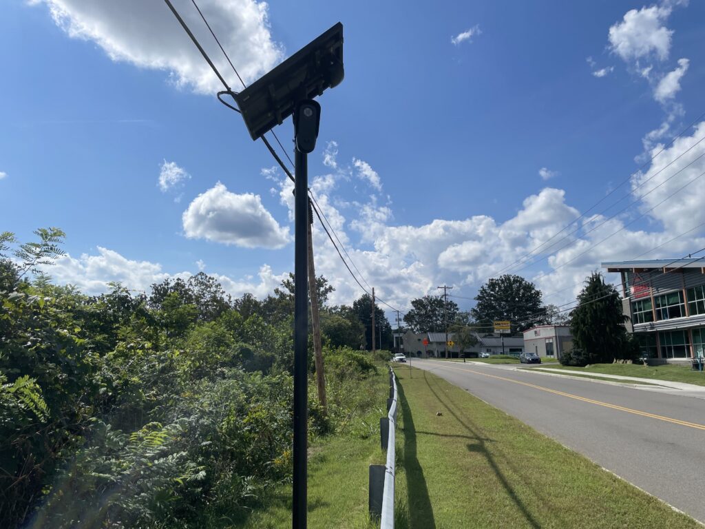 A Flock camera with a solar panel next to a guard rail.
