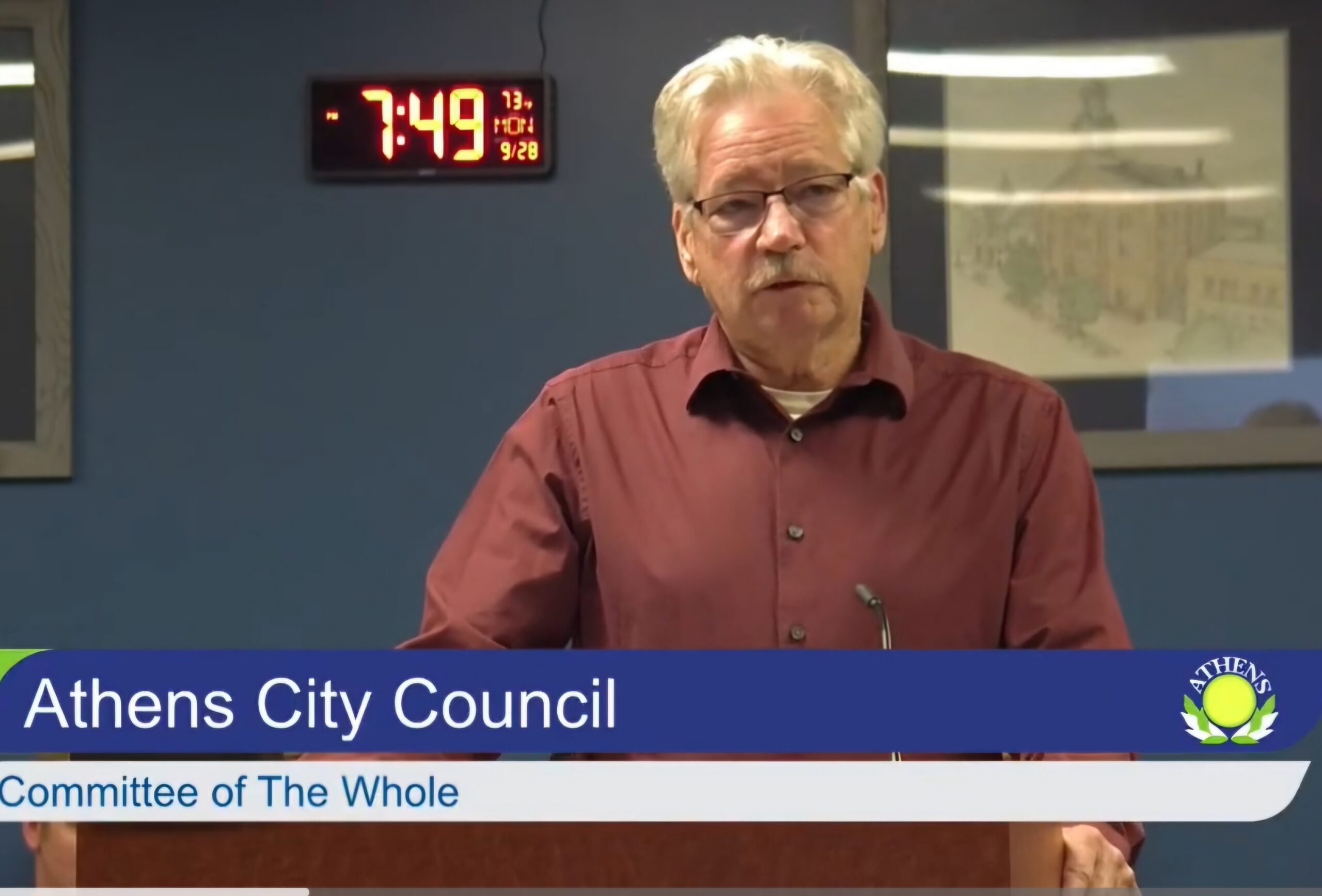 White man with white hair and glasses wearing a red shirt standing at a podium.