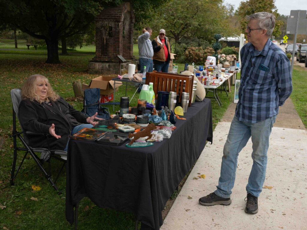 A woman, who is selling items from behind a table, talks to a man standing across from her.